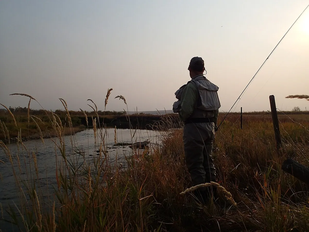 A fly angler in waders stands in tall grass at the edge of a river at dawn, his rod raised against a hazy sky.