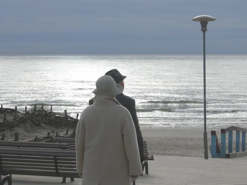 Seen from behind, an older couple in winter coats walks along a seaside promenade, the sea silver in the light.
