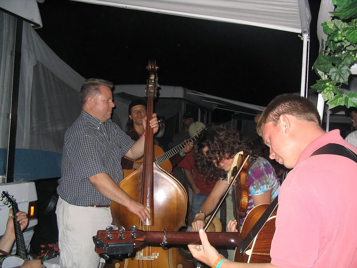At night under a tent awning, a man plays an upright bass beside fiddle and guitar players gathered close in a jam.