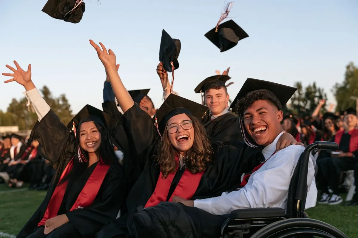 Graduates in black gowns laugh and cheer as caps fly overhead; one graduate sits in a wheelchair among friends.