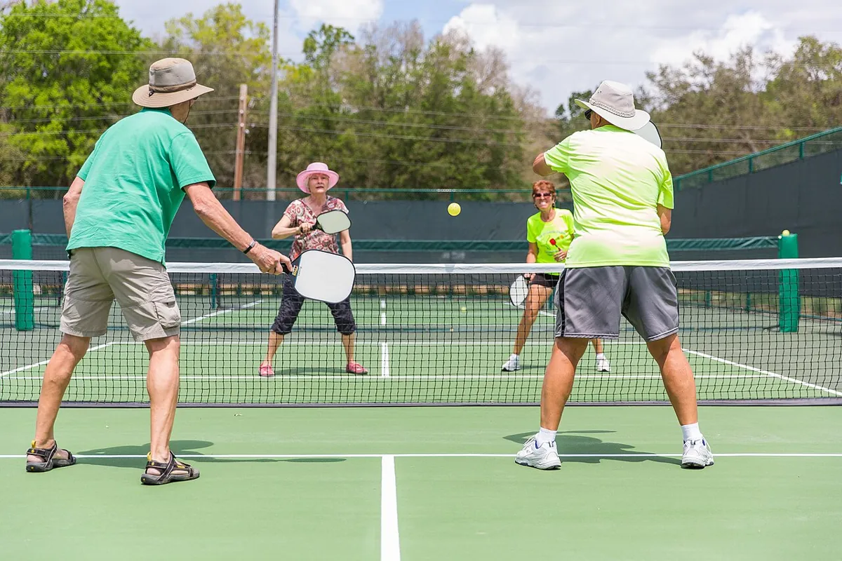 Four players in sun hats play pickleball on a green outdoor court on a bright day.
