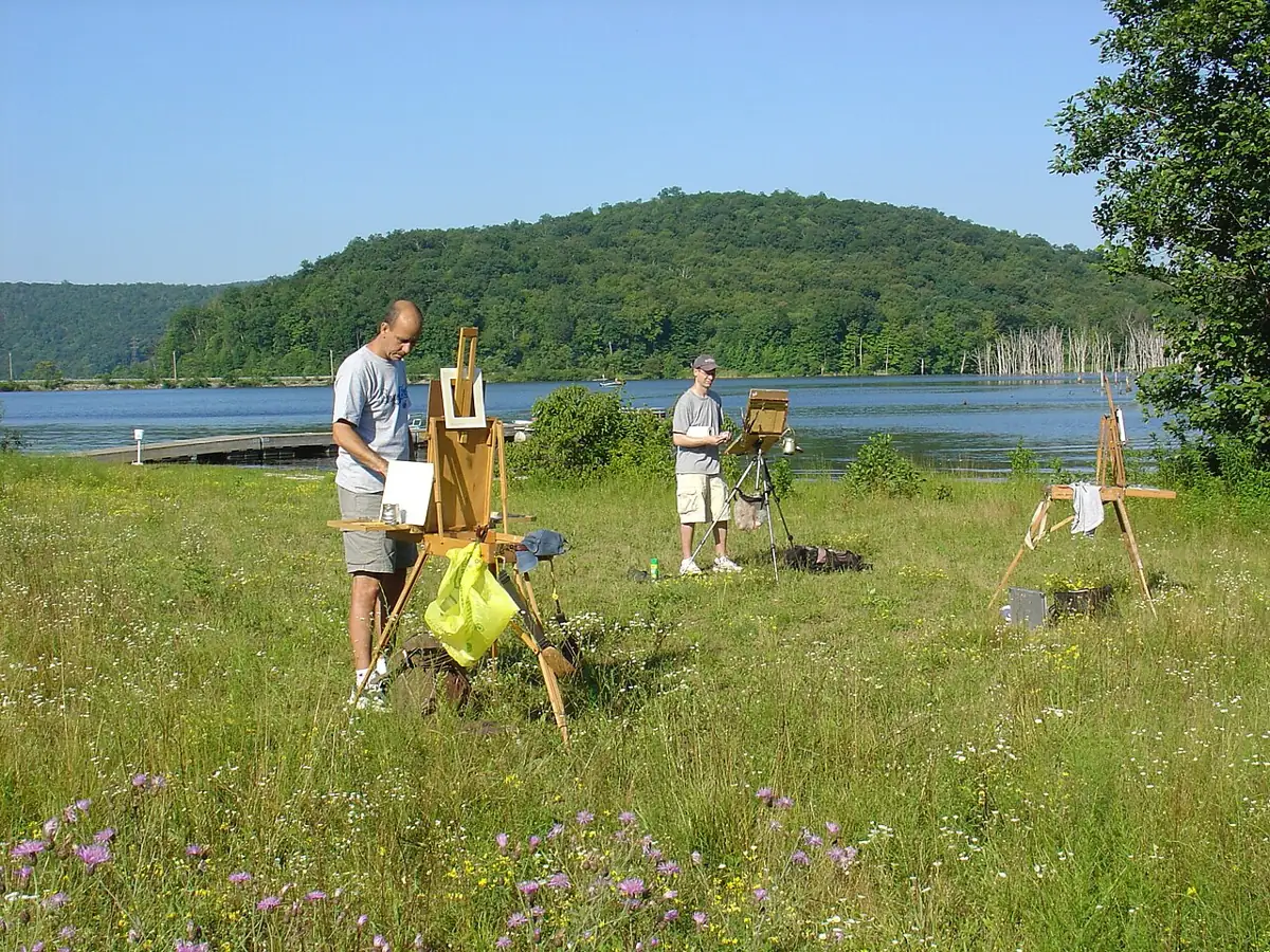 Painters stand at easels in a meadow of wildflowers beside a lake, a wooded hill across the water.