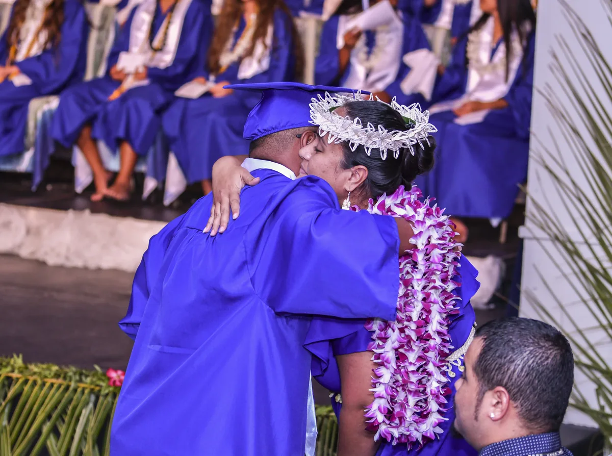 A graduate in a blue cap and gown hugs a woman wearing a flower crown and leis at a high school graduation.
