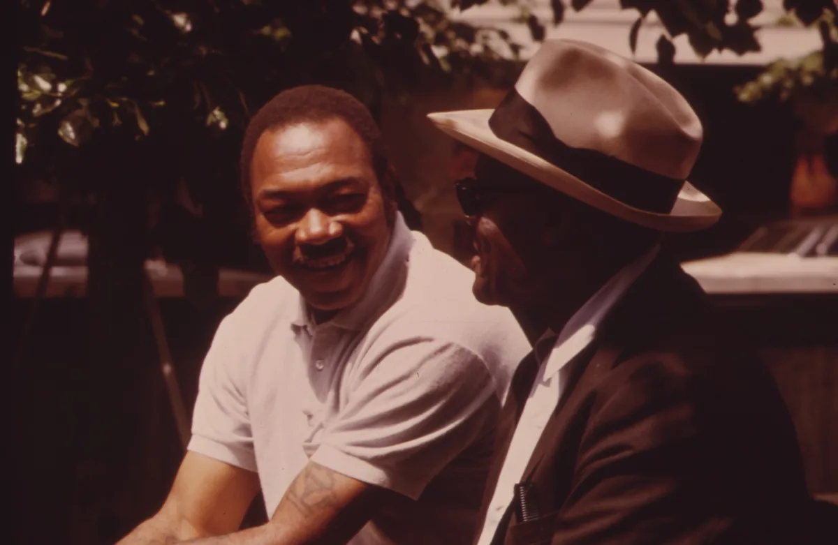 Two men talk and smile together outdoors in a city square.