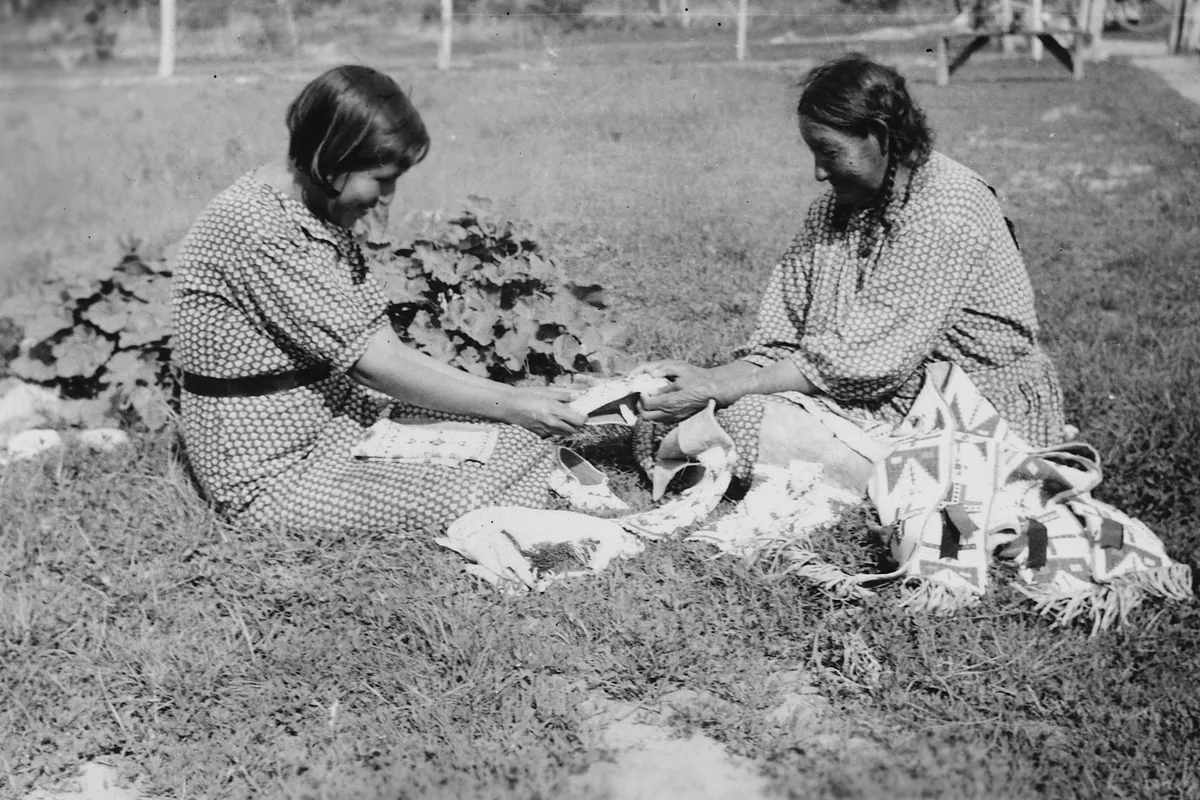 A grandmother teaches her granddaughter beadwork as the two sit on the grass.