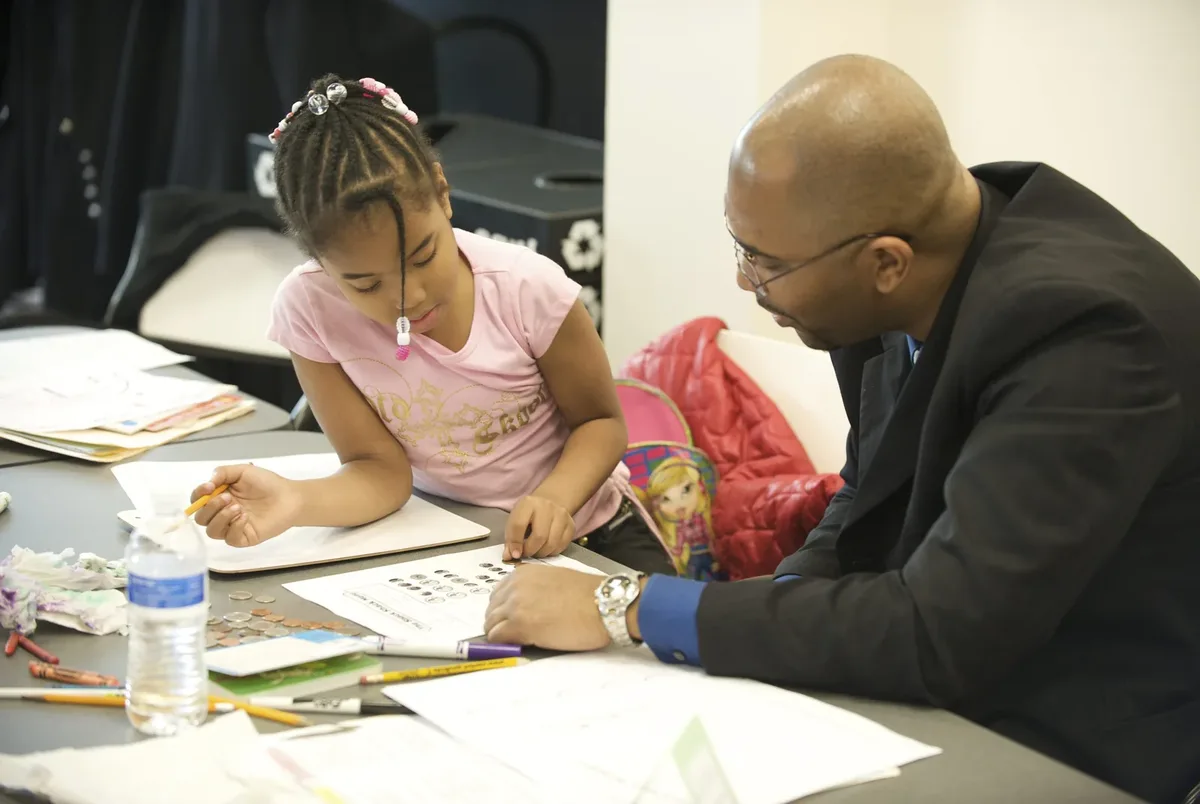 A man leans in beside a girl at a table as she works on a homework sheet, markers and papers spread around them.